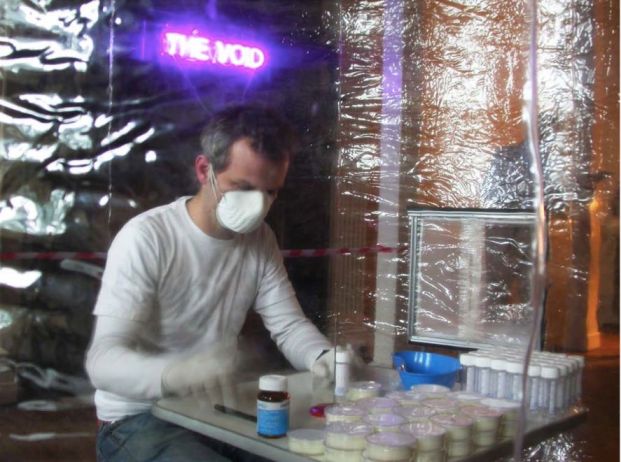 An artist in a respiratory air mask sits in a plastic tent. Test tubes and bottles on a table in front of him.
