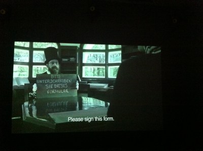 Film still (black and white) of a man holding a blackboard on which some German words are chalked, the translation is "You have to sign this form"