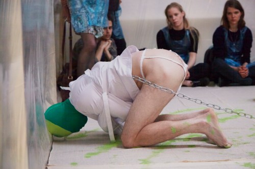 Main in straightjacket and rubber mask smears green paint with his head while people look on