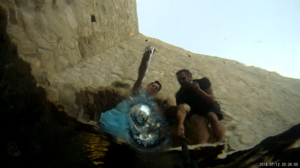 Looking upwards from beneath water, two people can be seen looking down into the water at what could be a silvery bubble that partially obscures them.