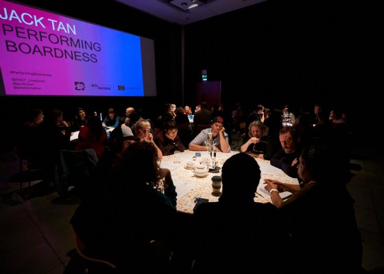 People gather round several lit round tables in a darkened room (about 8 people on each table). They look deeply engaged in conversation. Behind them a large screen has the caption: JACK TAN. PERFORMING BOARDNESS.