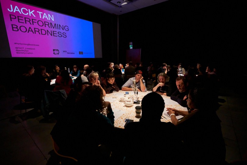 People gather round several lit round tables in a darkened room (about 8 people on each table). They look deeply engaged in conversation. Behind them a large screen has the caption: JACK TAN. PERFORMING BOARDNESS.